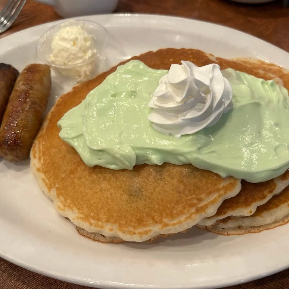 Stack of pancakes topped with green cream and whipped cream, served with two sausage links and butter.